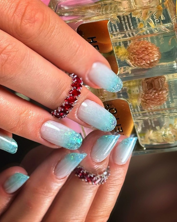 Close-up of beautifully manicured aqua blue nails with a glitter finish, showcasing elegant rings and a soft background.