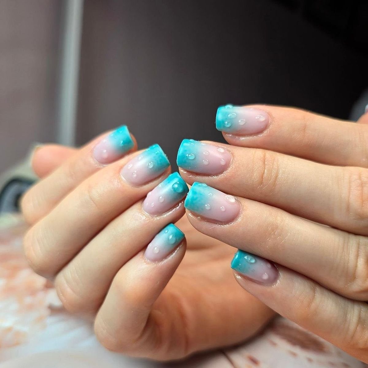 Two hands showcasing aqua and pink gradient nails adorned with tiny drops in a softly lit setting.