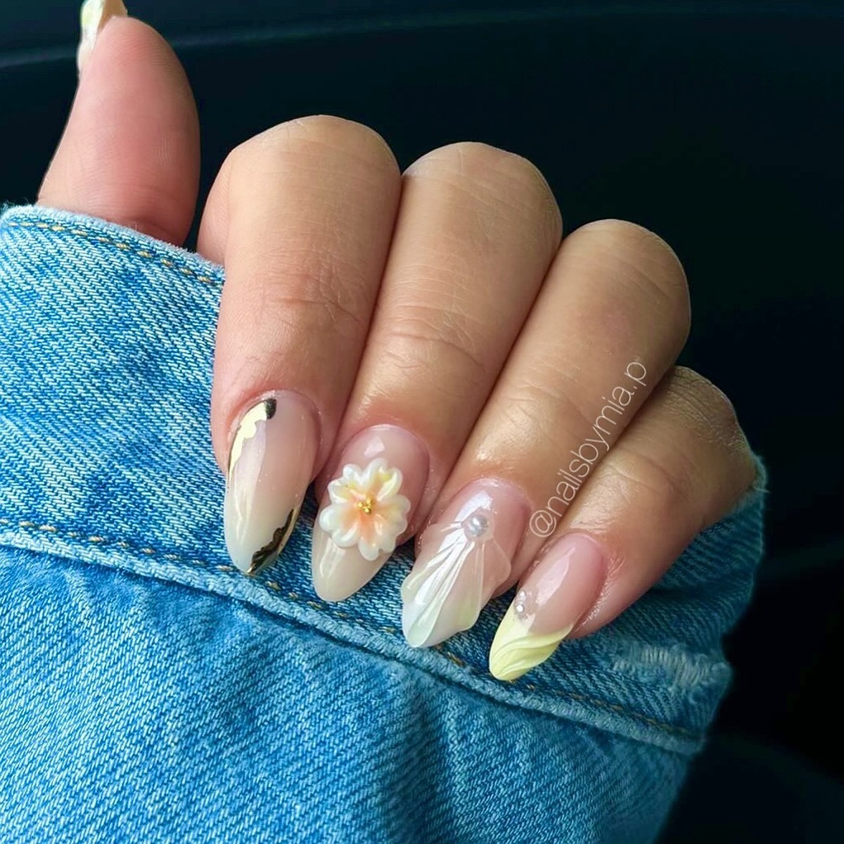 A hand featuring elegant beige floral nail art with long nails against a denim backdrop.