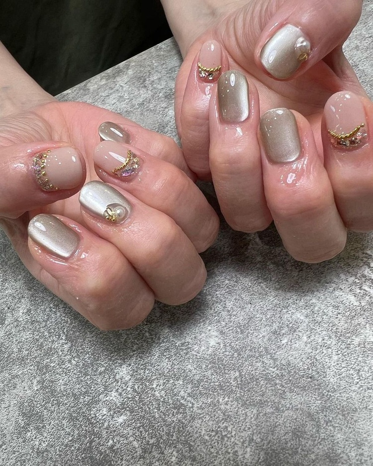 Two hands displaying elegant beige nails adorned with gold glitter and decorative embellishments.