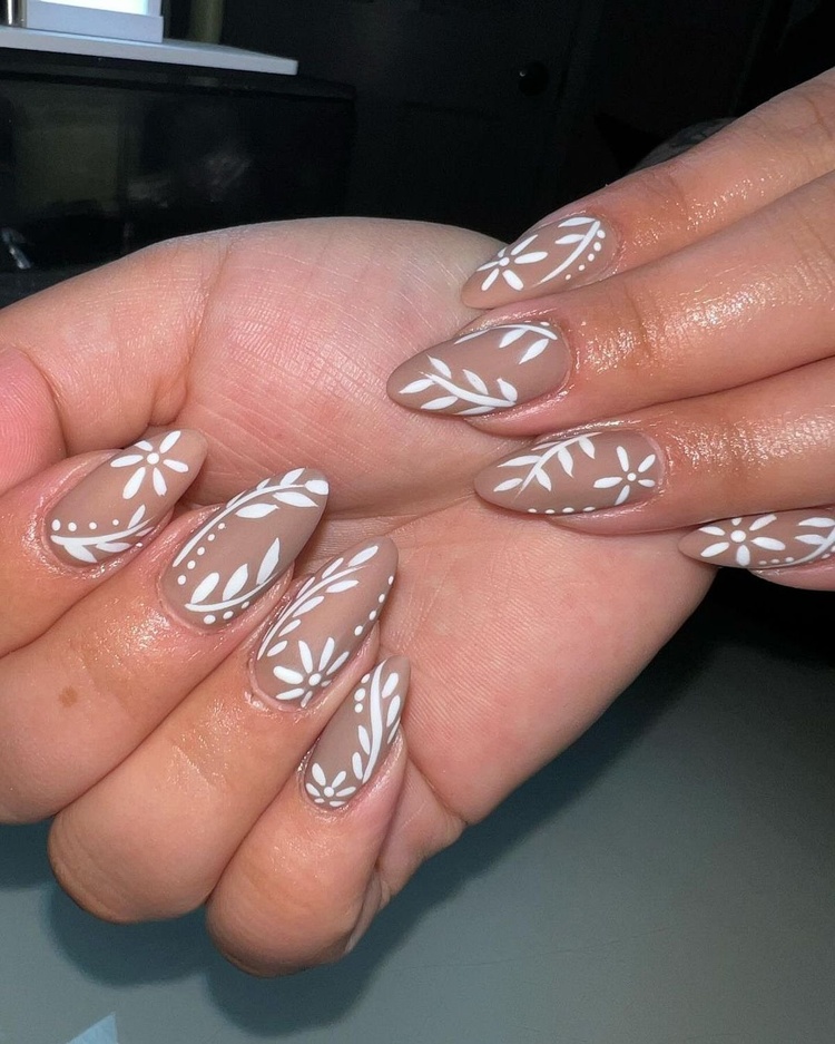 Close-up of elegant beige nails adorned with intricate white floral designs, showcasing a modern manicure style.