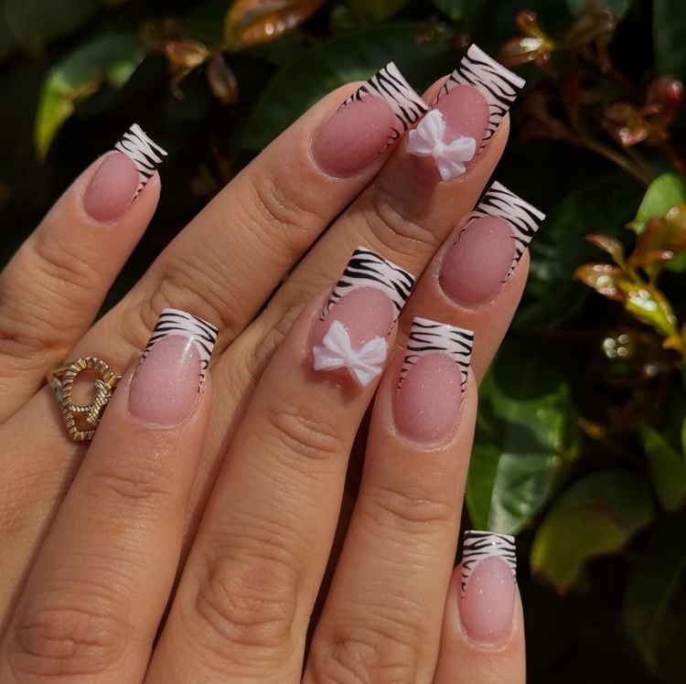 This image showcases stylish zebra print nails with white bows on a set of hands, surrounded by lush green leaves.